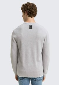 Grauer Strickpullover mit langen Ärmeln, Rundhalsausschnitt und strukturiertem Finish. Etikett mit sichtbarem Markennamen auf der Rückseite. Trägt man zu blauen Jeans.