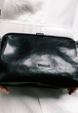 Sac à main Balagan en cuir noir tenu par deux mains sur un fond de tissu blanc.