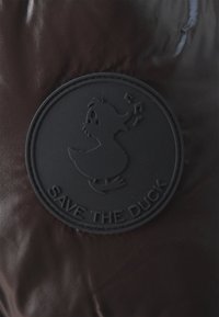 Bruin glanzend pufferstof bevat een opvallende zwarte rubberen patch met een eend afgebeeld, omringd door muzieknoten en de tekst "RED DE EEND."