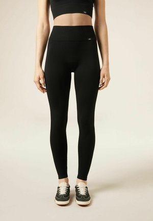 Frau trägt schwarze, hoch taillierte Leggings und ein passendes bauchfreies Oberteil mit schwarz-weißen Turnschuhen, steht vor einem einfarbigen Hintergrund.