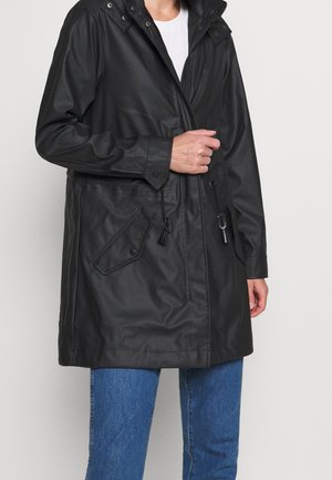 Person iført en sort vandtæt jakke med hætte, med klaplommer og snoretræk over en hvid skjorte og blå jeans.