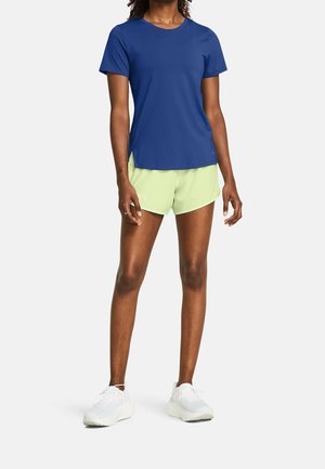 Mujer con una camiseta deportiva azul de manga corta, pantalones cortos para correr de color verde claro y zapatillas blancas, de pie sobre un fondo sencillo.