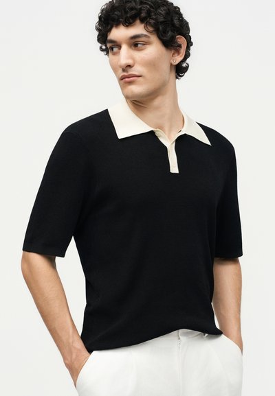 Homme aux cheveux bouclés portant un polo noir à manches courtes avec un col blanc et un pantalon blanc, les mains dans les poches, regardant sur le côté.