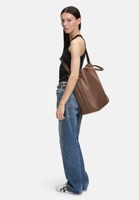 Bruine leren tote bag met een zachte, gestructureerde vorm; voorzien van lange schouderbanden en een bovenhandvat. Draagt door een persoon in casual kleding.