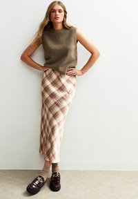 Ärmelloses olivfarbenes Strickoberteil kombiniert mit einem cremefarbenen Karorock in Rost- und Beigetönen. Dunkle Loafers mit goldenen Schnallsdetails.