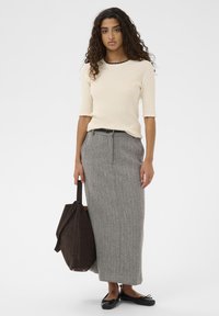 Femme aux cheveux bouclés portant un haut crème à côtes, une jupe grise en chevrons, une ceinture noire, des ballerines noires et portant un grand sac à main brun foncé.