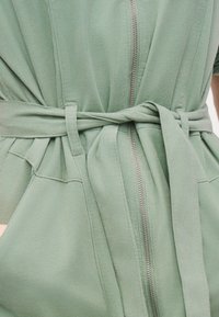 Μαλακό πράσινο ρουχιστικό από κοτλέ με φερμουάρ στο μπροστινό μέρος και ζώνη από το ίδιο ύφασμα δεμένη στη μέση πάνω από μεγάλες πλαϊνές τσέπες.