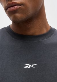 Κοντινό πλάνο στο κάτω μέρος του προσώπου και το λαιμό ενός άνδρα που φοράει σκούρο γκρι μπλουζάκι με στρογγυλή λαιμόκοψη και μικρό λευκό λογότυπο Reebok στο στήθος.