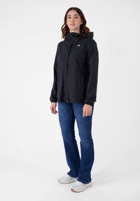 Giacca nera impermeabile con zip frontale, polsini elastici, cappuccio regolabile e logo. Indossata sopra jeans blu e sneakers bianche.