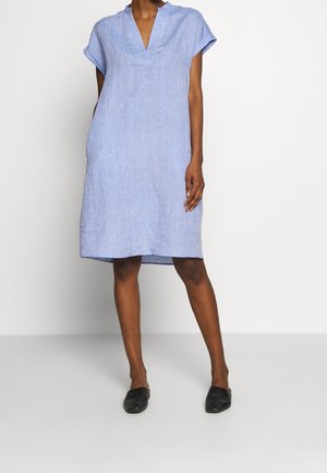 Mujer vestida con un vestido azul claro de manga corta hasta la rodilla, con escote en V y zapatos negros sin cordones, de pie contra una pared blanca.