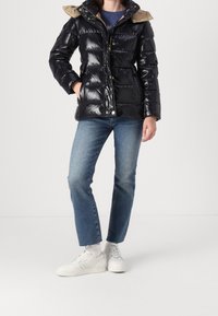 Gepolsterte schwarze Steppjacke mit glänzendem Finish, beigem Kragen, Frontreißverschluss und Druckknöpfen, kombiniert mit blauen Jeans und weißen Turnschuhen.