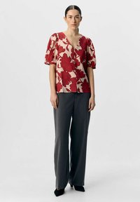 Blusa de estampado floral rojo con mangas abullonadas, escote en V profundo y cierre de botones, combinada con pantalones de pierna ancha grises y zapatos negros de punta.