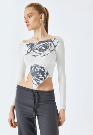Biały prążkowany crop top z czarnym graficznym wzorem róży, odkryty dekolt i postrzępiony dół. W zestawie z szarymi dresami dla kontrastu.