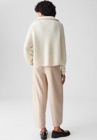 Maglione lavorato all'uncinetto in crema con un ampio colletto e texture a coste, abbinato a pantaloni beige a gamba stretta e scarpe scure.