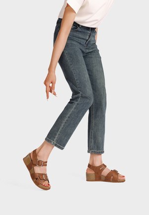 Persona che indossa jeans blu corti e sandali con zeppa marroni con doppie fibbie, in piedi con un piede leggermente sollevato.