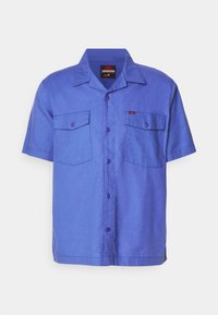 Kortärmad, knäppskjorta i ljusblått tyg. Har två bröstfickor, en spetsig krage och en liten röd logotagg.