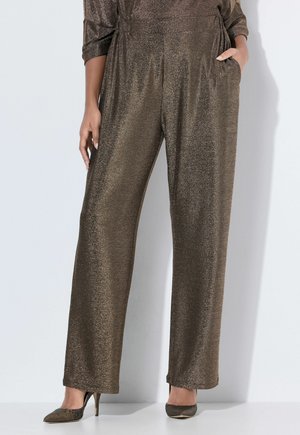 Vrouw draagt losse, glanzende bronzen wijd uitlopende broek en bijpassende hoge hakken, één hand in de zak, staand tegen een lichte achtergrond.