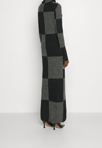 Lång, figursydd klänning i mörkgrå och svart rutmönster, tillverkad i stickad material, med hög krage och långa ärmar. Höga klackskor synliga.