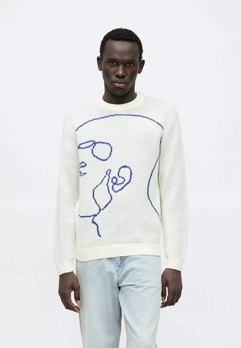 Gebreide trui in crème met een blauwe lijntekening van een gezicht. Voorzien van een ronde hals en een gestructureerde weefstructuur. Gecombineerd met lichtblauwe jeans.