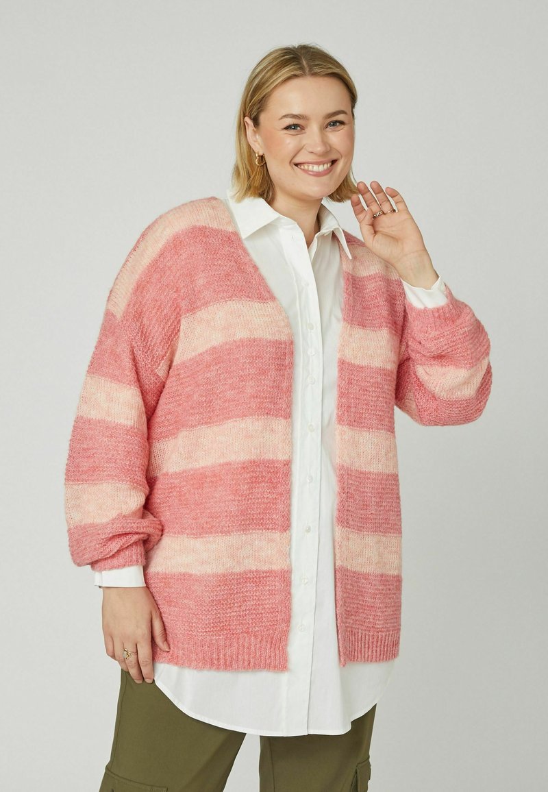 Femme souriante portant un cardigan rose et rose clair à rayures par-dessus une chemise blanche à boutons et un pantalon vert olive, levant une main près de son visage.