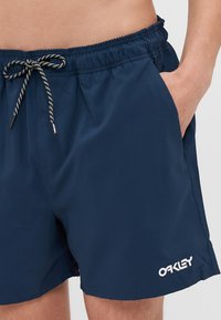 Marineblå svømshorts laget av lett materiale med elastisk midjebånd, snor og sidelommer. Har hvit logo brodering.