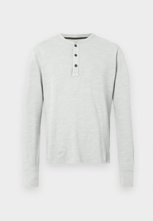 Svijetlosiva dugosleeve henley majica od mekog materijala, sa četiri dugmeta duž plavka i rebrastim manžetama. Glatka tekstura.