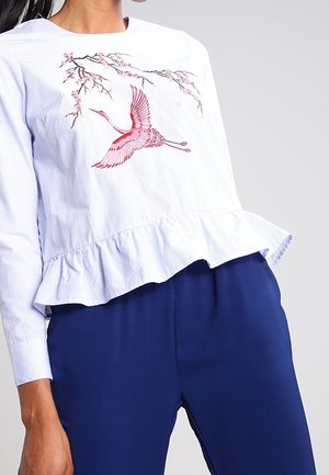 Biała bluzka z falbanką u dołu, zdobiona haftem z różowym żurawiem i gałązką kwitnącej wiśni, w zestawieniu z ciemnoniebieskimi spodniami.