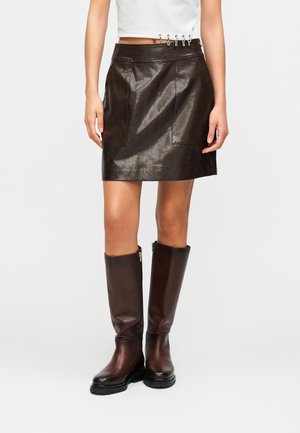 Gonna marrone in finta pelle con design aderente, caratterizzata da tasche frontali e dettagli decorativi in metallo sulla cintura; abbinata a stivali marroni al ginocchio.