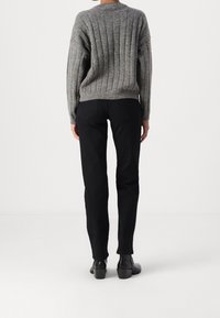 Person standing with back facing forward, wearing a gray ribbed sweater, black pants, and black heeled ankle boots on a white background.