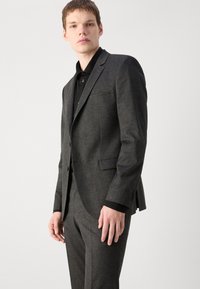 Grijze wollen kostuum met opstaande revers, eenzijdig gesloten ontwerp en twee voorzakken. Zwarte shirt eronder met knoopdetails.