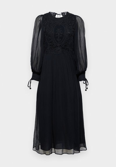 Černé šaty s průhlednými dlouhými rukávy. Podrobný krajkový korzet a výstřih. Splývavá sukně s pásky na zápěstích a strukturovanou tkaninou.