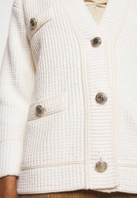 Kardigan o kremowej teksturze z dużymi złotymi dekoracyjnymi guzikami z przodu, na rękawach i klapie kieszeni, noszony na beżowym swetrze.