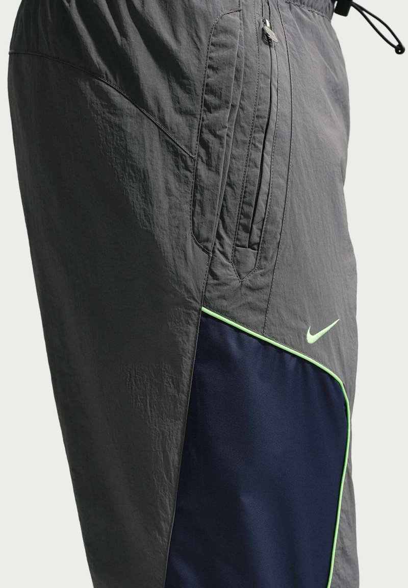 Grijze en marineblauwe sportbroek met een neongroen Nike-logo en bies, zijritszak en trekkoord in de tailleband, getoond van dichtbij.