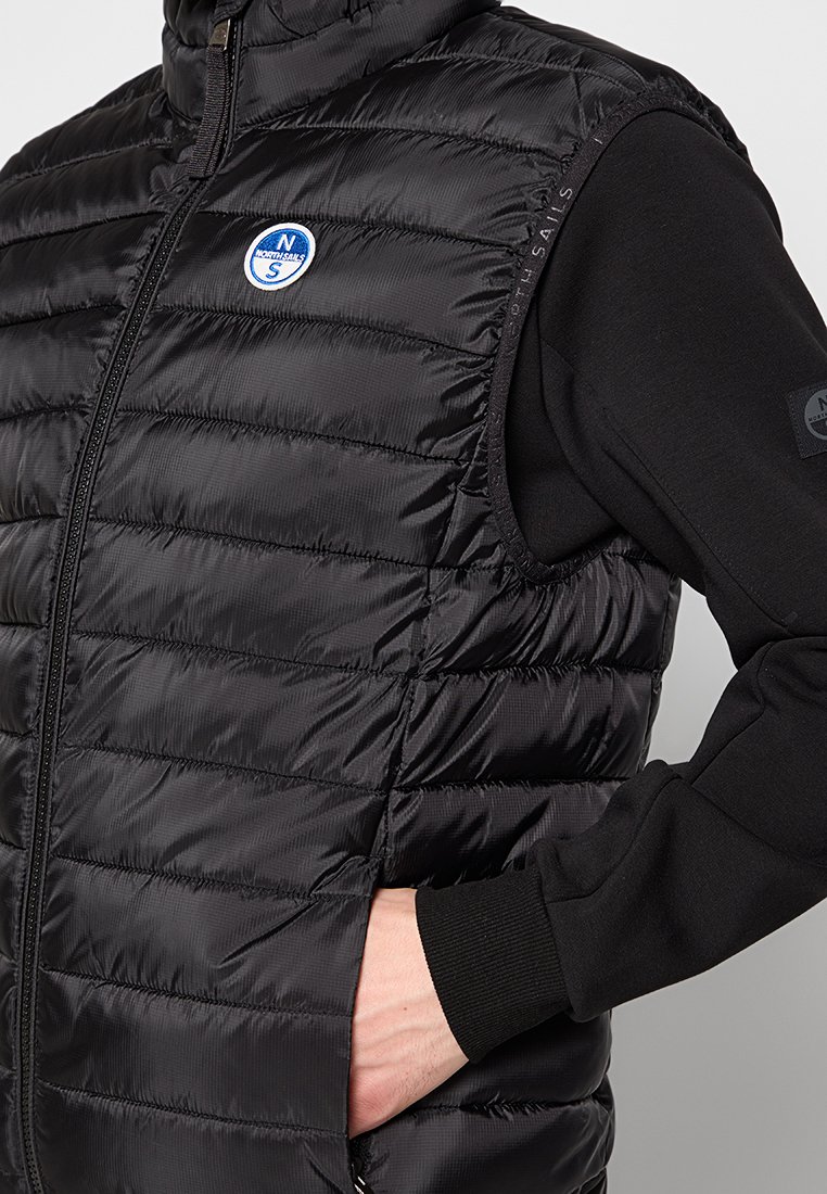 Gilet nero in piuma con zip frontale, dotato di cuciture orizzontali, un badge con logo rotondo e aperture per le braccia. Indossato sopra una maglietta nera a maniche lunghe.
