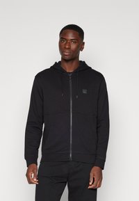 Svart zip-up hoodie med dragsko i huvan, med texturerad söm och en liten logotypapplikation på bröstet. Släta tyget och figurnära design.