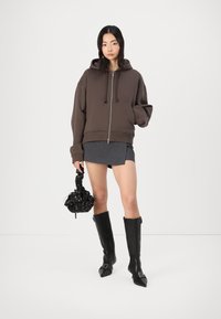 Sudadera con cremallera marrón, con una gran capucha, llevada sobre una mini falda gris. Botas negras hasta la rodilla, sosteniendo un pequeño bolso negro con flores.