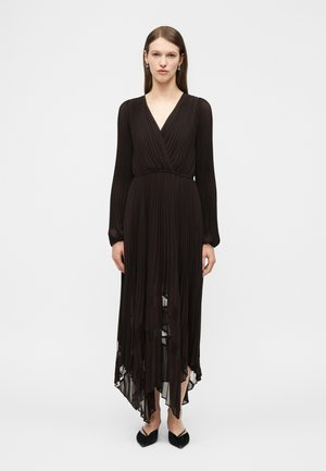 Kobieta ubrana w czarną plisowaną sukienkę z długim rękawem, dekoltem w serek i asymetrycznym dołem, w połączeniu z czarnymi butami na spiczastym nosku, stojąca na białym tle.