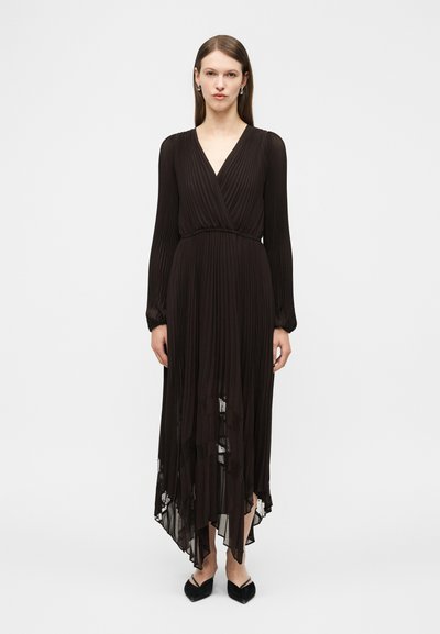 Femme portant une robe plissée noire à manches longues avec un col en V et un ourlet asymétrique, associée à des chaussures noires à bout pointu, debout sur un fond blanc.