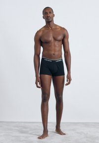 Black boxer briefs with a white logo waistband, shown on a standing male model against a plain light background.
