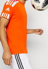 Oranžové sportovní tričko s bílými pruhovanými akcenty na rukávech. Černobílý fotbalový míč je držen blízko trička. Bílé šortky s černými pruhy.