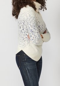 Maglione color crema con collo alto, con motivi a macchie blu, polsini a coste e un orlo posteriore leggermente più lungo, abbinato a jeans blu scuro.