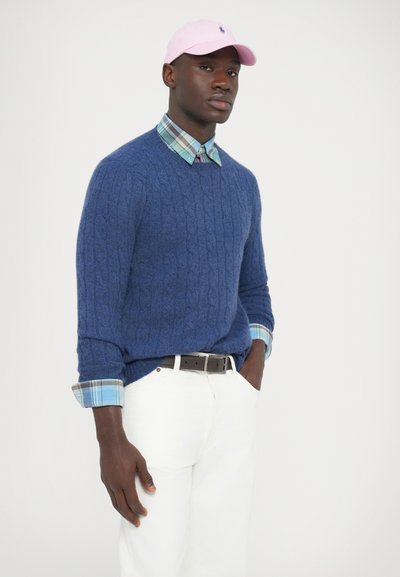 Kék kötött pulóver kockás inggel, fehér nadrággal és rózsaszín sapkával. Figyelemre méltó textúrák és rétegzett minták láthatók.