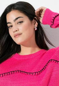 Suéter de punto de color rosa brillante con un acabado texturizado y detalles de costura negra. Pequeño pendiente de aro de plata con una superficie brillante.