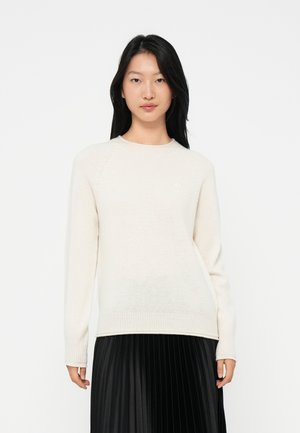 Donna con lunghi capelli neri che indossa un maglione a maglia crema e una gonna plissettata nera, in piedi davanti a uno sfondo bianco semplice.