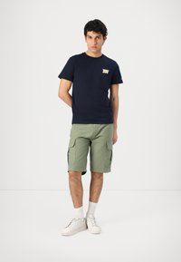 T-shirt di cotone blu navy con un piccolo logo sul petto, abbinato a pantaloni cargo verde oliva con tasche laterali e vestibilità rilassata. Sneakers bianche.
