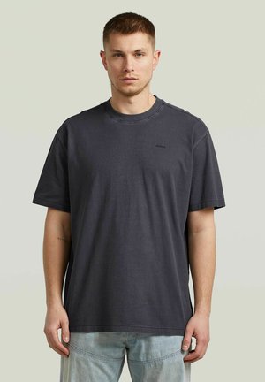 Młody mężczyzna o krótkich włosach, ubrany w gładki, ciemnoszary, luźny T-shirt i jasnoniebieskie dżinsy, stojący na neutralnym tle.