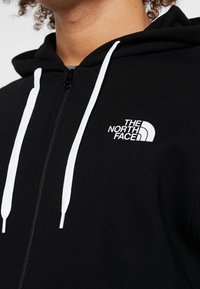 Felpa nera con zip e coulisse bianche, e logo. Il tessuto ha una texture liscia. Presenta un collo alto e un design aderente.