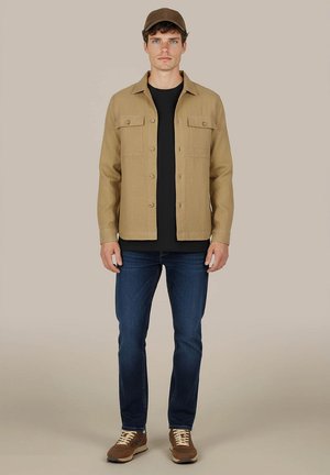 Mann mit brauner Kappe, beiger Knopfleistenjacke, schwarzem Hemd, blauen Jeans und braunen Sneakern steht vor einem einfarbigen Hintergrund.