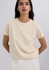 Camiseta de manga corta en color blanco roto, confeccionada en suave algodón, con cuello redondo y un pequeño logo de calavera impreso en el pecho. Textura suave, corte relajado.