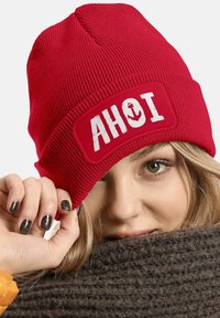 Red knit beanie with a rectangular patch featuring the word "AHOI" and an anchor design. Ribbed texture with a snug fit.